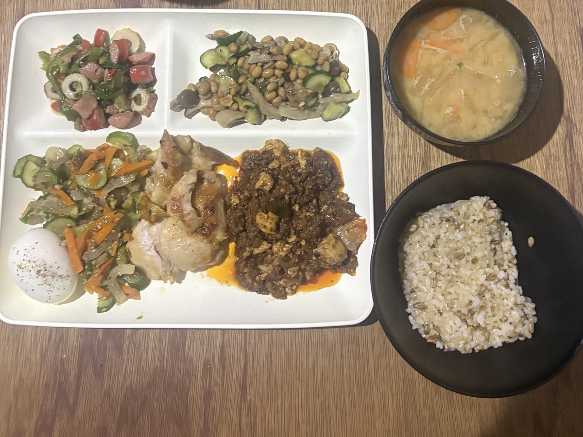 仕切りプレートに盛った鶏もも肉のソテーと麻婆豆腐、ゆで卵、きゅうりと人参の酢の物、ピーマンとちくわとトマトの和え物、しめじと大豆ときゅうりの和え物。具だくさんの味噌汁と玄米ご飯