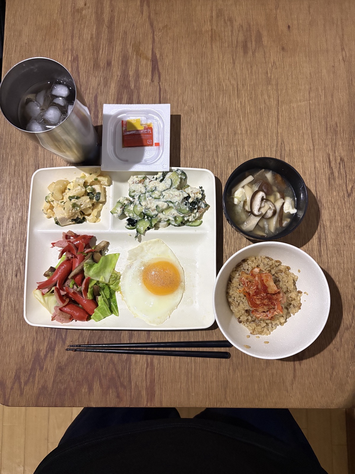 仕切りプレートに盛った豆腐と卵の炒め、目玉焼き、きゅうりの和えもの、魚肉ソーセージとレタスのサラダ。きのこと油揚げの味噌汁と、玄米にキムチをのせた茶碗、納豆つき