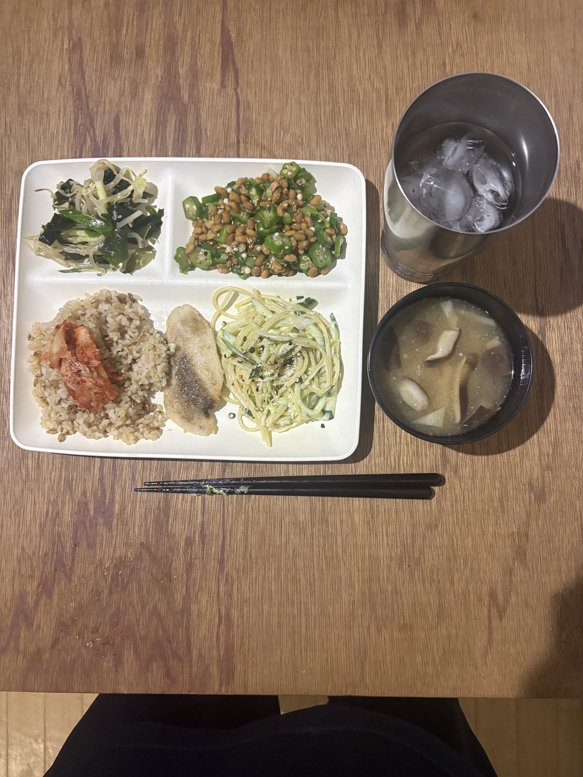 仕切りプレートに盛った白身魚のムニエル、スパゲッティサラダ、わかめときゅうりともやしの酢の物、オクラ納豆。きのこの味噌汁と、玄米にキムチをのせたごはん、氷水のグラス